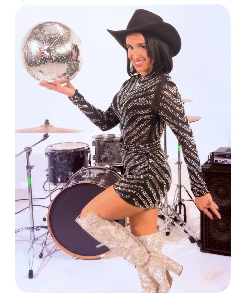 Jackie in a sparkling zebra-print mini dress and black cowboy hat, holding a mirror-ball disco ball in front of a black drum kit and bass amp.