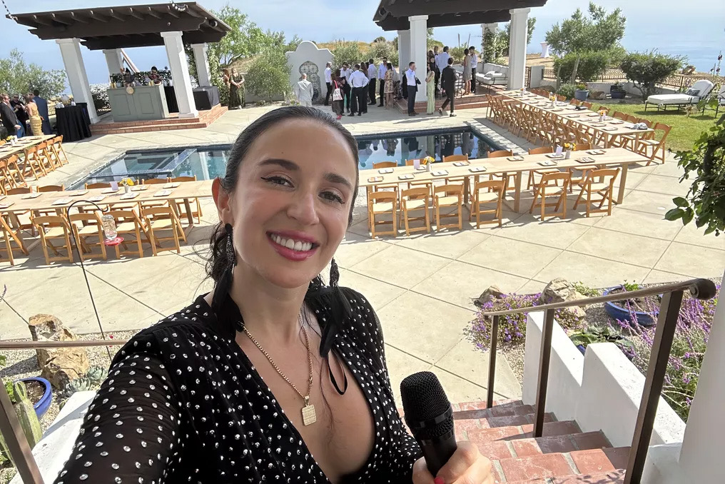 Jackie holding a wireless mic in front of an oceanview pool with rows of wedding chairs facing the altar.